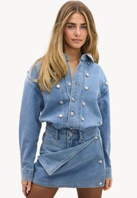 Femme aux longs cheveux blonds portant une veste en jean et une mini-jupe en jean assortie avec des détails de boutons sur les deux vêtements.