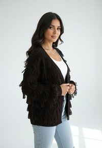 Ciemnobrązowy dziergany kardigan z ozdobnymi frędzlami, otwartym przodem i ściągaczami na mankietach, w połączeniu z białym topem i jasnoniebieskimi dżinsami.