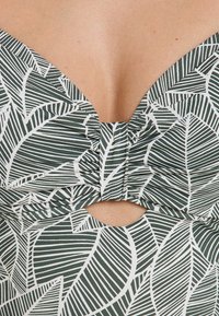 Maillot de bain à motifs de feuilles avec un décolleté en V profond, un devant froncé et un détail découpé. Les couleurs incluent le vert foncé et le blanc, avec un tissu lisse et extensible.