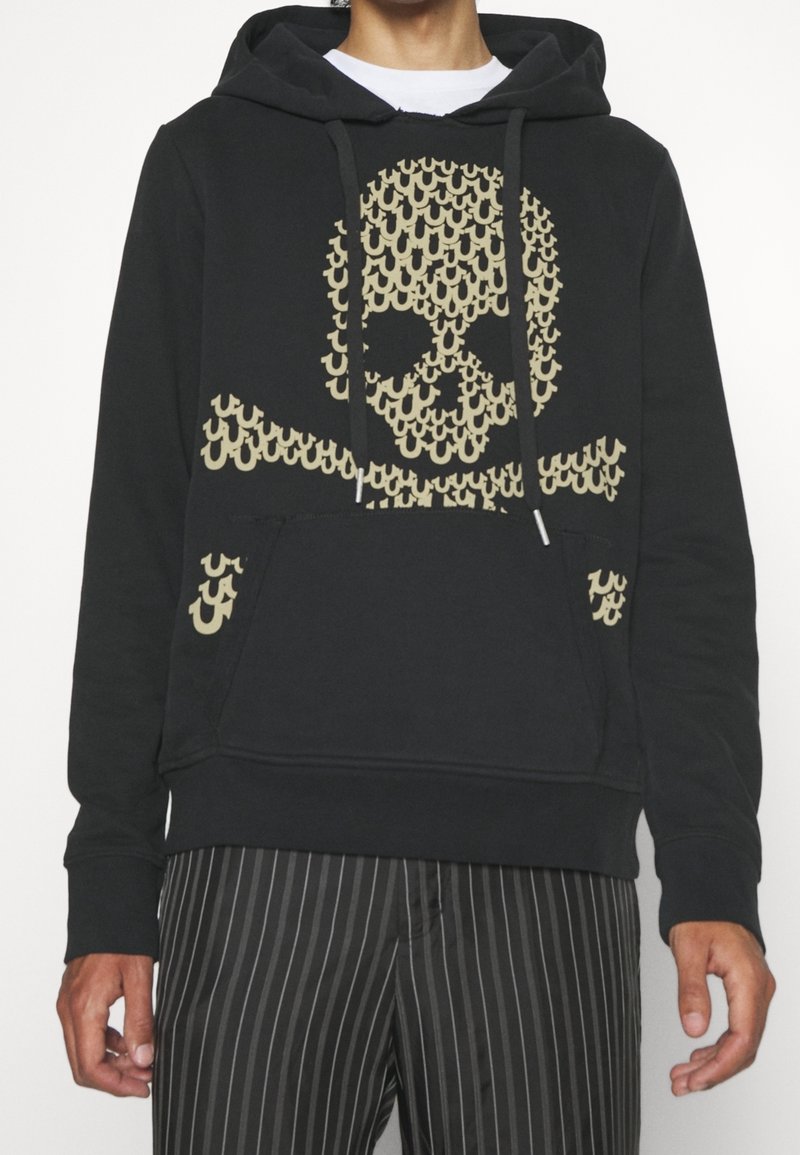 Personne portant un sweat à capuche noir avec un motif beige de tête de mort et os croisés composé de petites formes répétées, associé à un pantalon noir à fines rayures.