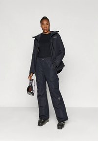 Žena v čiernom lyžiarskom oblečení a topánkach, ktorá drží čierno-biele lyžiarske okuliare pred svetlým jednoliatym pozadím.