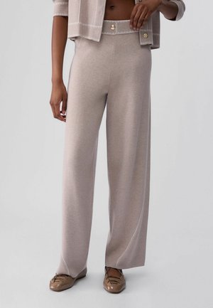 Persona che indossa pantaloni beige a gamba larga in maglia con bottoni dorati e cardigan beige coordinato, abbinati a mocassini marroni lucidi.