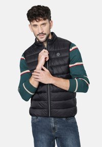 Chaleco acolchado negro con cuello alto, cierre frontal y parche de logo. Usado sobre una camiseta de manga larga de rayas verdes, combinado con jeans azules.