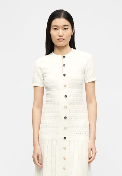 Νεαρή γυναίκα με ίσια σκούρα μαλλιά φοράει ένα κοντομάνικο, λευκό φόρεμα με κουμπιά και υφασμένους μοτίβους, στέκεται μπροστά σε απλό φωτεινό φόντο.
