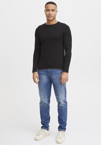 Svart langermet tettsittende topp med rund hals, kombinert med lyseblå jeans og hvite sneakers, som står foran en ensfarget bakgrunn.