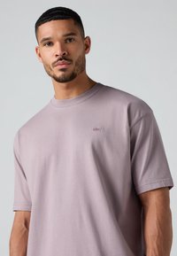 T-shirt en coton violet clair avec un col rond, des manches courtes et un petit logo sur le côté gauche de la poitrine. Texture lisse.