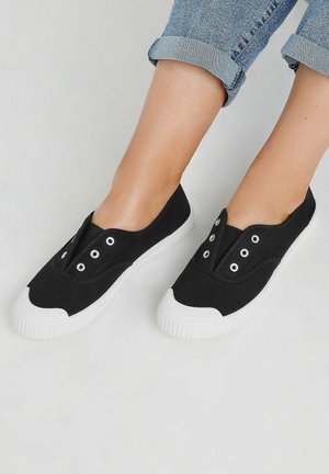 Des chaussures slip-on noires en toile avec une semelle en caoutchouc blanche, dotées de plusieurs œillets et d'un design de cape de orteil incurvé unique.