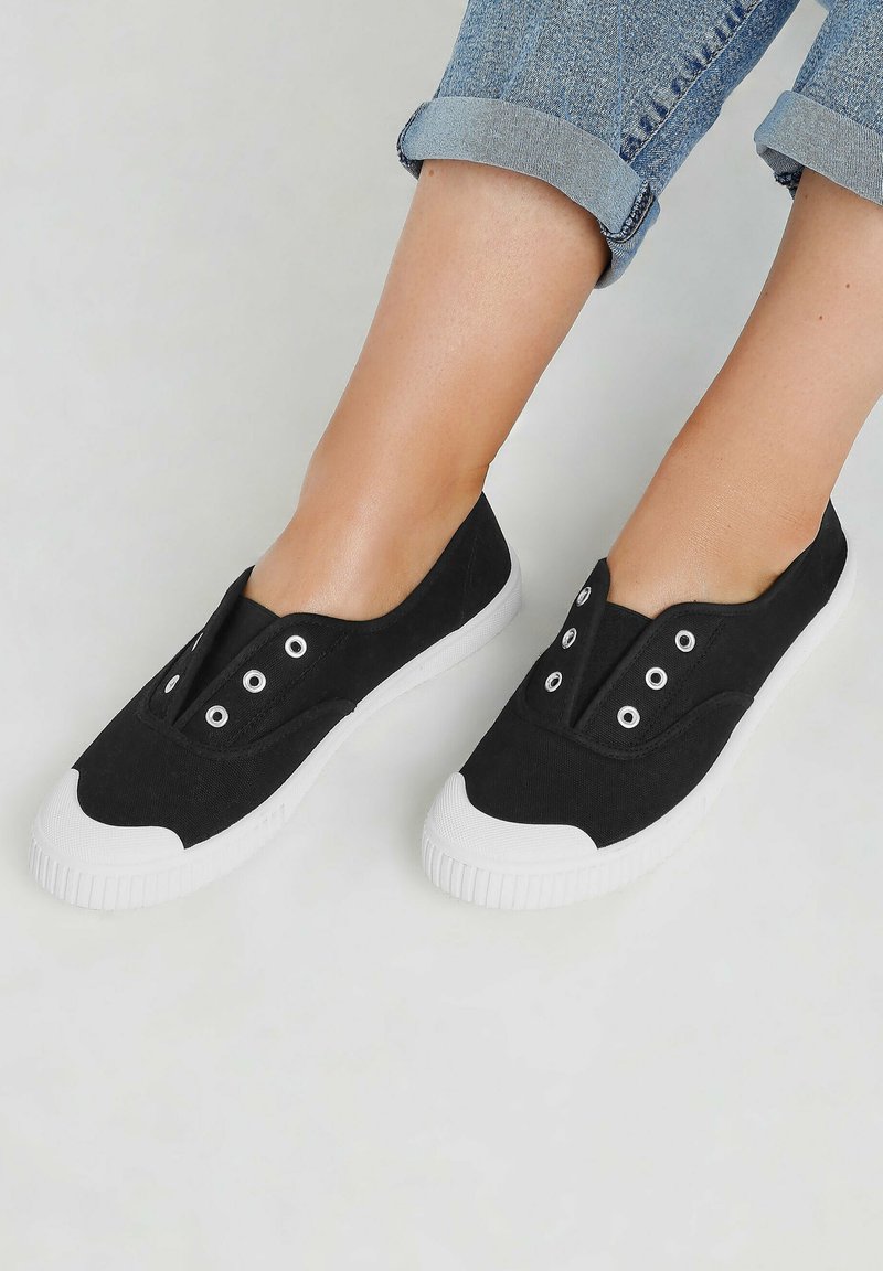 Scarpe slip-on nere in tela con suola in gomma bianca, caratterizzate da molteplici occhielli e un design unico della punta curvata.