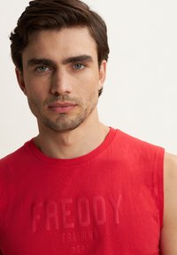 Joven con cabello corto castaño y barba ligera, vestido con una camiseta sin mangas roja que tiene el texto "FREDDY TRAINING DEPT".