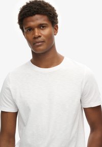 Camiseta de algodón blanca con un clásico cuello redondo y mangas cortas, con un acabado suave y texturizado. Diseño minimalista, sin adornos.