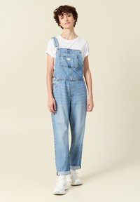 Pantalones de mezclilla de color azul claro con tirantes ajustables, bolsillo frontal y puños enrollados. Combinados con una camiseta blanca y zapatillas blancas.