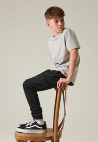 Tricou gri deschis cu mâneci scurte, asortat cu pantaloni de trening negri, așezat pe un scaun de lemn. Adidași negri cu șireturi albe și șosete scurte.