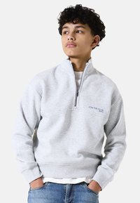 Sudadera gris con cremallera y cuello alto, y puños acanalados. Presenta "ÚNETE AL CLUB" bordado en navy. Combinada con jeans de denim azul.