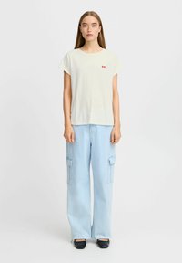 Lys cremefarvet t-shirt med korte ærmer og en rød sløjfe broderet på venstre bryst, parret med højhalsede lyseblå cargo bukser og sorte sko.