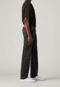 Hombre de pie de lado, vistiendo una camiseta negra, jeans oscuros y zapatillas blancas, contra un fondo beige liso.