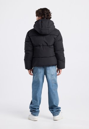 Chaqueta de plumas negra con capucha, con un diseño acolchado. Combinada con unos vaqueros holgados de color azul claro y unas zapatillas blancas.