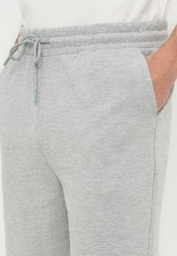 Κοντινό πλάνο ενός ατόμου που φορά ανοιχτό γκρι φόρμες με κορδόνι στη μέση και το ένα χέρι στην πλαϊνή τσέπη.