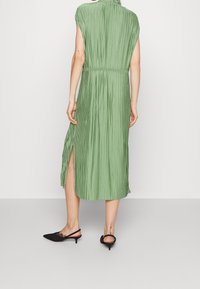 Grünes plissiertes Midikleid mit elastischem Bund, kurzen Ärmeln und seitlichen Schlitzen. Kombiniert mit schwarzen spitzen Slingback-Pumps.