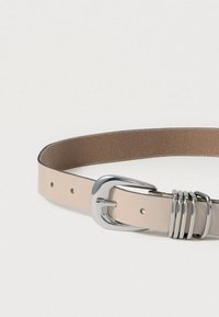 Ceinture en cuir beige avec une boucle en métal argenté brillant et des anneaux décoratifs, présentant une texture lisse et des bords soigneusement cousus.