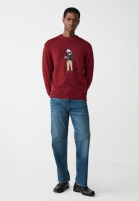 Bordowy sweter dziany z ribbowanym dekoltem, z haftowanym jeżem w garniturze, w połączeniu z niebieskimi dżinsami i czarnymi loafersami.