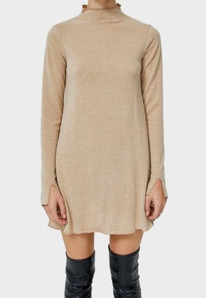 Beżowa dzianinowa sukienka na długi rękaw z wysokim kołnierzem, marszczonym dekoltem, rozkloszowanym dołem i bocznymi rozcięciami, noszona z czarnymi kozakami sięgającymi kolan.