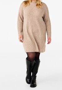 Strickkleid in hellem Beige, mit vertikalen Rippen und geometrischen Mustern. Kombiniert mit schwarzen kniehohen Stiefeln.