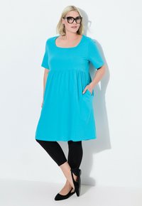 Vestido corto turquesa de manga corta con escote cuadrado y cintura fruncida, con bolsillos laterales, combinado con pantalones legging negros y zapatos negros.