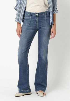 Frau trägt mittelblaue Bootcut-Jeans mit aufgesetzten Fronttaschen, ein gestreiftes Shirt und beige offene flache Schuhe.