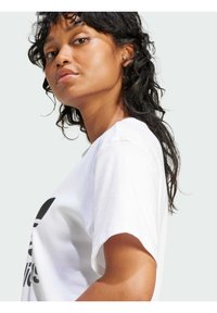 Camiseta blanca de algodón con mangas cortas, que presenta una gran impresión gráfica negra en la parte frontal. El modelo tiene el cabello ondulado y un fondo neutro.