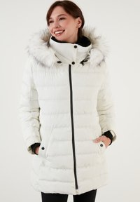 Veste blanche matelassée avec une capuche en fausse fourrure, accents noirs, fermeture éclair sur le devant, poches latérales et un bas légèrement évasé. Texture lisse.