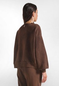 Sweatshirt oversize marrone realizzato in tessuto morbido, con spalle cadenti, polsini a costine e una sottile cucitura decorativa nel retro del collo.