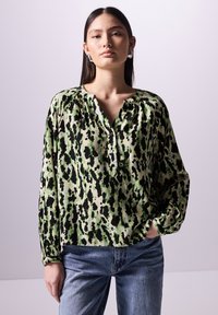 Grün und beige bedruckte Bluse mit schwarzen Punkten, mit V-Ausschnitt und langen Ärmeln, aus leichtem Stoff. Kombiniert mit blauen Jeans.