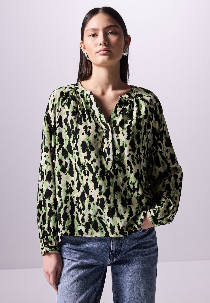 Grün und beige bedruckte Bluse mit schwarzen Punkten, mit V-Ausschnitt und langen Ärmeln, aus leichtem Stoff. Kombiniert mit blauen Jeans.