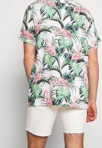 Camicia in tessuto a maniche corte con stampa tropicale caratterizzata da fenicotteri rosa e foglie verdi su sfondo bianco, abbinata a pantaloni corti in denim bianchi.