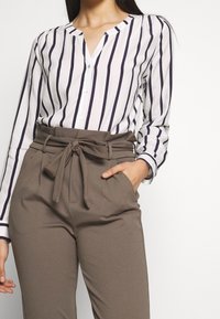 Blouse rayée blanche et marine, avec encolure en V, boutonnière sur le devant et manches longues. Associée à un pantalon marron taille haute avec ceinture nouée.