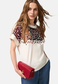 Camiseta blanca de manga corta con un diseño de lentejuelas en degradado en rosa y negro. Un clutch de cuero rojo con un clasp dorado complementa el atuendo.