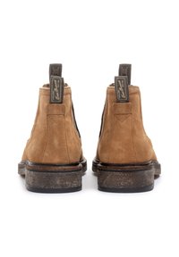 Paire de bottines Chelsea en daim marron avec des languettes de tirage noires et des semelles texturées marron foncé, vues de dos sur un fond blanc.