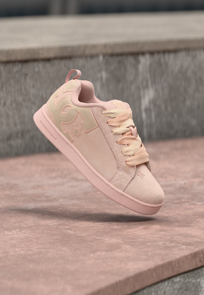 Baskets en daim rose clair avec des lacets épais, grand logo sur le côté et semelle en caoutchouc sur un fond de surface en pierre.