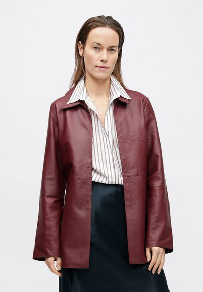 Γυναίκα που φοράει ένα μπορντό δερμάτινο μπουφάν πάνω από ένα λευκό ριγέ πουκάμισο και μια μαύρη φούστα, στέκεται μπροστά σε ένα απλό ανοιχτό φόντο.