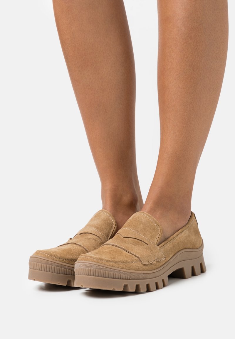 Chaussures à enfiler en daim couleur fauve, avec une semelle en caoutchouc rainurée, ornées de coutures décoratives et d'une finition lisse. Portées pieds nus.