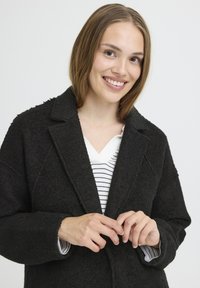 Cappotto nero in tessuto texturizzato con colletto a revers, caratterizzato da una vestibilità ampia e chiusura a bottoni, indossato sopra una maglietta a righe bianche.