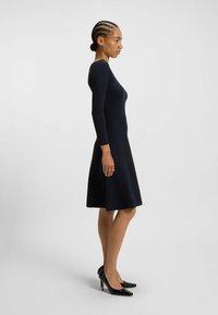 Robe longue de couleur marine à manches longues en tissu texturé, corsage ajusté, jupe évasée. Modèle associé à des chaussures à talons noirs, vue de profil.