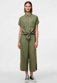Une combinaison vert olive à manches courtes, avec un devant boutonné, deux poches poitrine et une taille à nouer. Assortie à des baskets noires.