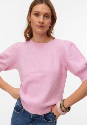 Femme aux cheveux châtain clair portant un pull à manches courtes rose clair, un jean bleu et des bracelets au poignet droit, debout les mains sur les hanches.
