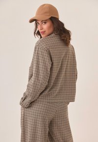 Mujer con un traje beige de diseño y una gorra a juego, mirando por encima del hombro contra un fondo claro y liso.
