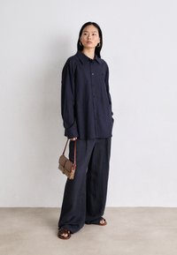 Camisa de botones azul marino con cuello, combinada con pantalones anchos azul marino. Los accesorios incluyen un bolso marrón con estampado y sandalias marrones.
