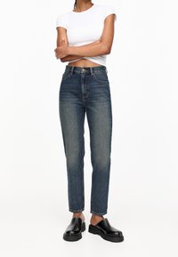 High-waisted blue jeans featuring a straight-leg cut, subtle fading, five pockets, and a classic button closure, paired with black slip-on shoes.