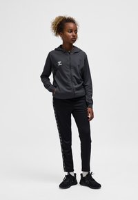 Junge Frau steht mit den Händen in den Taschen, trägt einen schwarzen Kapuzenpullover mit Reißverschluss, passende Jogginghosen mit Seitenstreifen, weiße Socken und schwarze Turnschuhe.