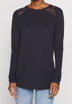 Kvinde iført en sort langærmet top med mønstrede meshpaneler på skuldrene og lyseblå jeans, stående mod en ensfarvet baggrund.