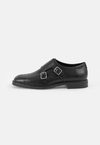 Des chaussures en cuir noir présentent un bout pointu, deux boucles en argent sur une sangle à travers le coup de pied, et une semelle en caoutchouc plate avec une légère texture.